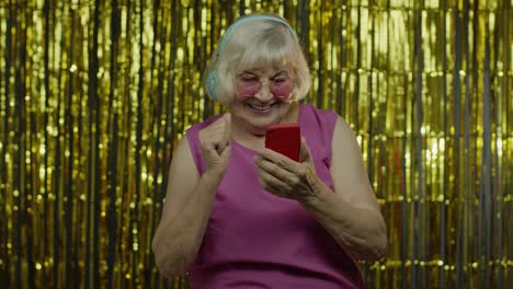 Stylish-happy-senior-old-woman-holding-cell-phone,-listening-to-music-in-headphones-and-dancing