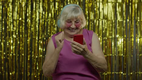 stylish happy senior old woman holding cell phone, listening to music in headphones and dancing