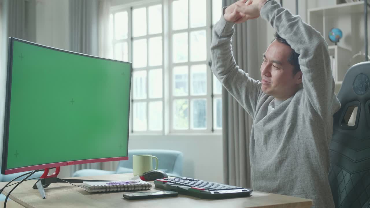 Asian Man In Long Sleeved T-Shirt With Green Screen Desktop Computer Stretching And Leaning Back While Working At Home.