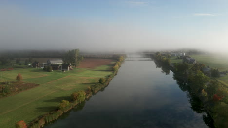 La-Niebla-Matutina-Se-Cierne-Sobre-Un-Río-Tranquilo-Y-Granjas-Verdes-Pacíficas-A-Lo-Largo-De-Las-Orillas