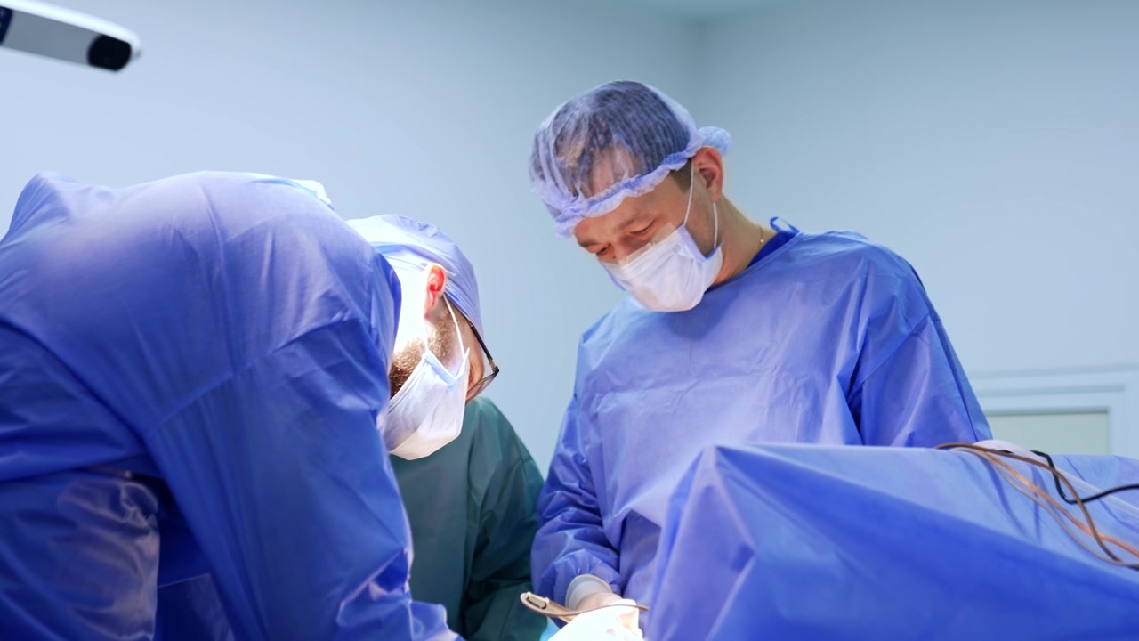 Neurochirurgo barbuto si china sul paziente per utilizzare uno strumento elettrico. Intervento neurochirurgico eseguito da un'équipe medica.