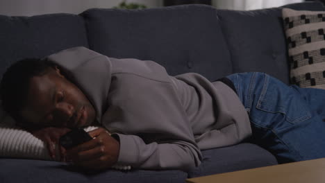 unhappy and depressed young man lying on sofa at home at night looking at mobile phone shaking his head