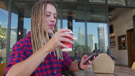 Mann-Gemischter-Abstammung-Mit-Dreadlocks-Sitzt-An-Einem-Tisch-Vor-Einem-Café,-Trinkt-Kaffee-Und-Benutzt-Ein-Smartphone