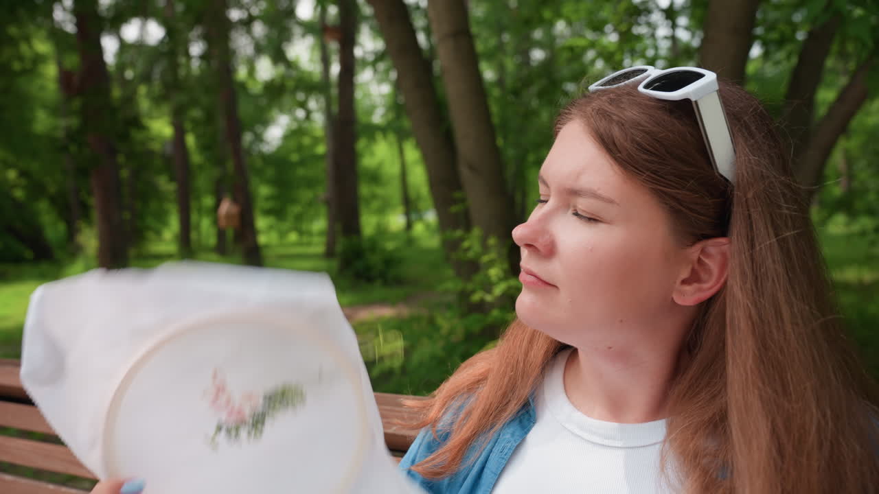 Mujer elegante sentada en un banco del parque abanicándose la cara en un día soleado, con los ojos entrecerrados y mirando a la distancia, gesto de alivio por el calor veraniego, gafas de sol blancas en la cabeza, camisa azul informal, con un bosque verde de fondo