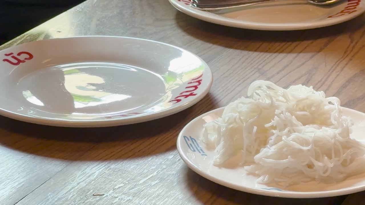 Hand uses fork and spoon to move rice noodles onto plate in soft natural lighting