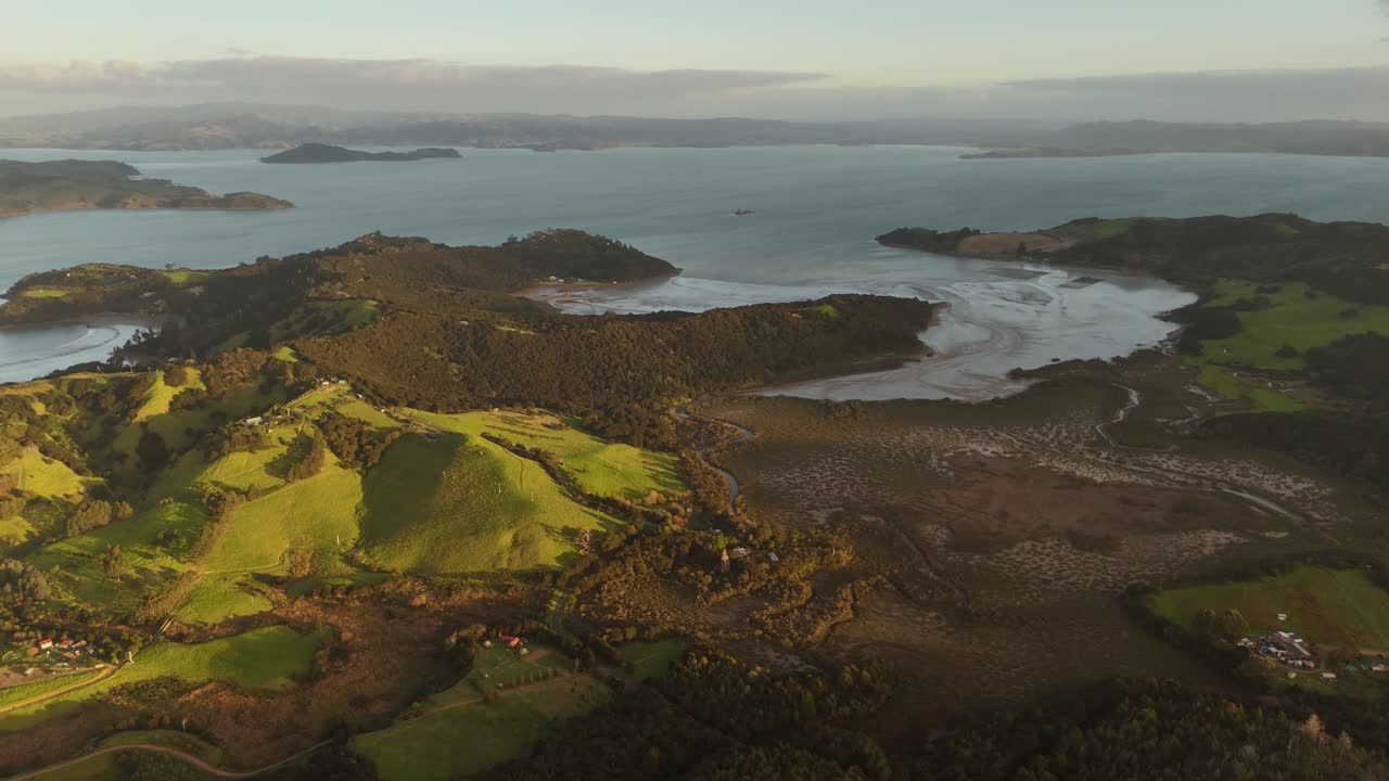 A stunning 4K 60fps aerial view of Waiheke Island at sunset. The drone glides over lush, rolling green hills and the pastoral coastline, bathed in beautiful golden hour light. A serene NZ landscape