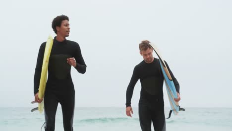 Vorderansicht-Von-Zwei-Männlichen-Surfern,-Die-Zusammen-Mit-Einem-Surfbrett-Am-Strand-Laufen,-4k