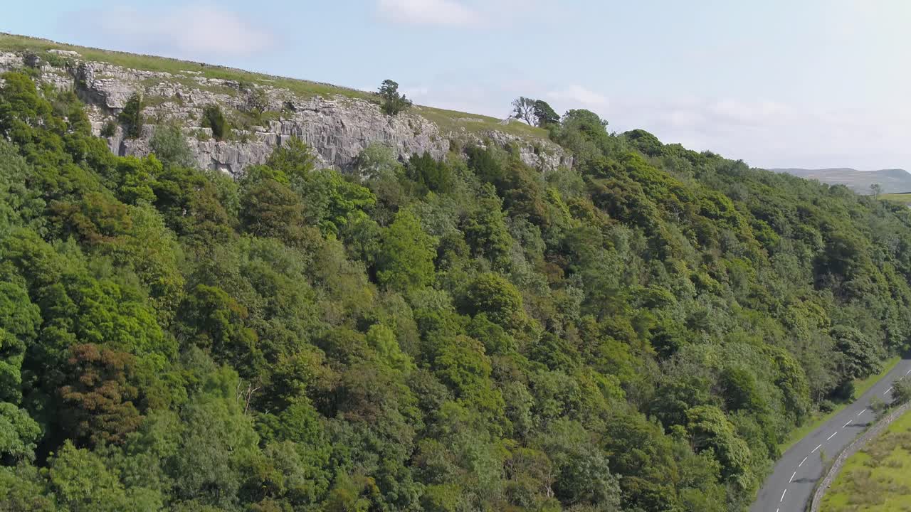 imágenes de drones de una carretera rural que sube por un acantilado montañoso bordeado de árboles que revela el campo de yorkshire, tierras de cultivo y campos, paredes de piedra seca con un paisaje montañoso en la distancia