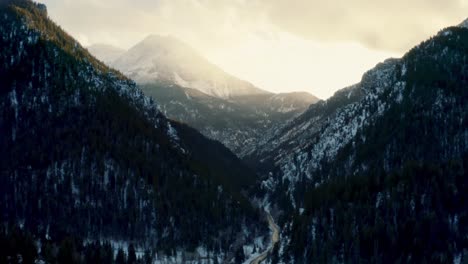 Rising-aerial-drone-shot-of-a-winter-landscape-of-Mount-Timpanogos-in-the-background-surrounded-by-a-pine-tree-forest-during-sunset-from-the-frozen-Tibble-Fork-Reservoir-in-American-Fork-Canyon,-Utah