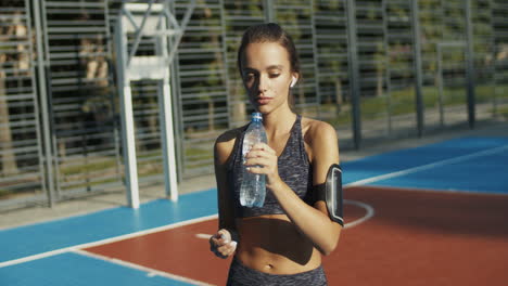 Sportliche-Frau,-Die-An-Einem-Außenplatz-Steht,-Sich-Ausruht-Und-Nach-Dem-Training-Kaltes-Wasser-Trinkt