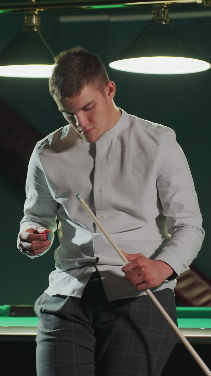 Giovane uomo in camicia bianca e pantaloni grigi seduto su un tavolo da biliardo verde, che tiene in mano una stecca e applica con attenzione una copertura rossa con espressione concentrata. Le luci dall'alto illuminano l'ambiente