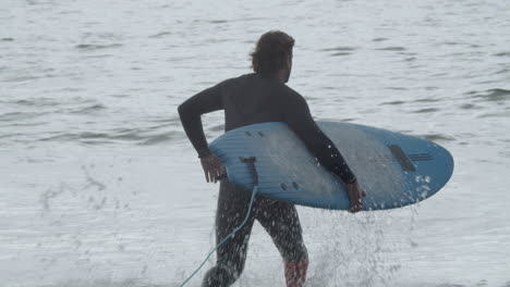 vista trasera de un hombre con pierna artificial en traje de neopreno entrando en el océano y acostado en la tabla de surf