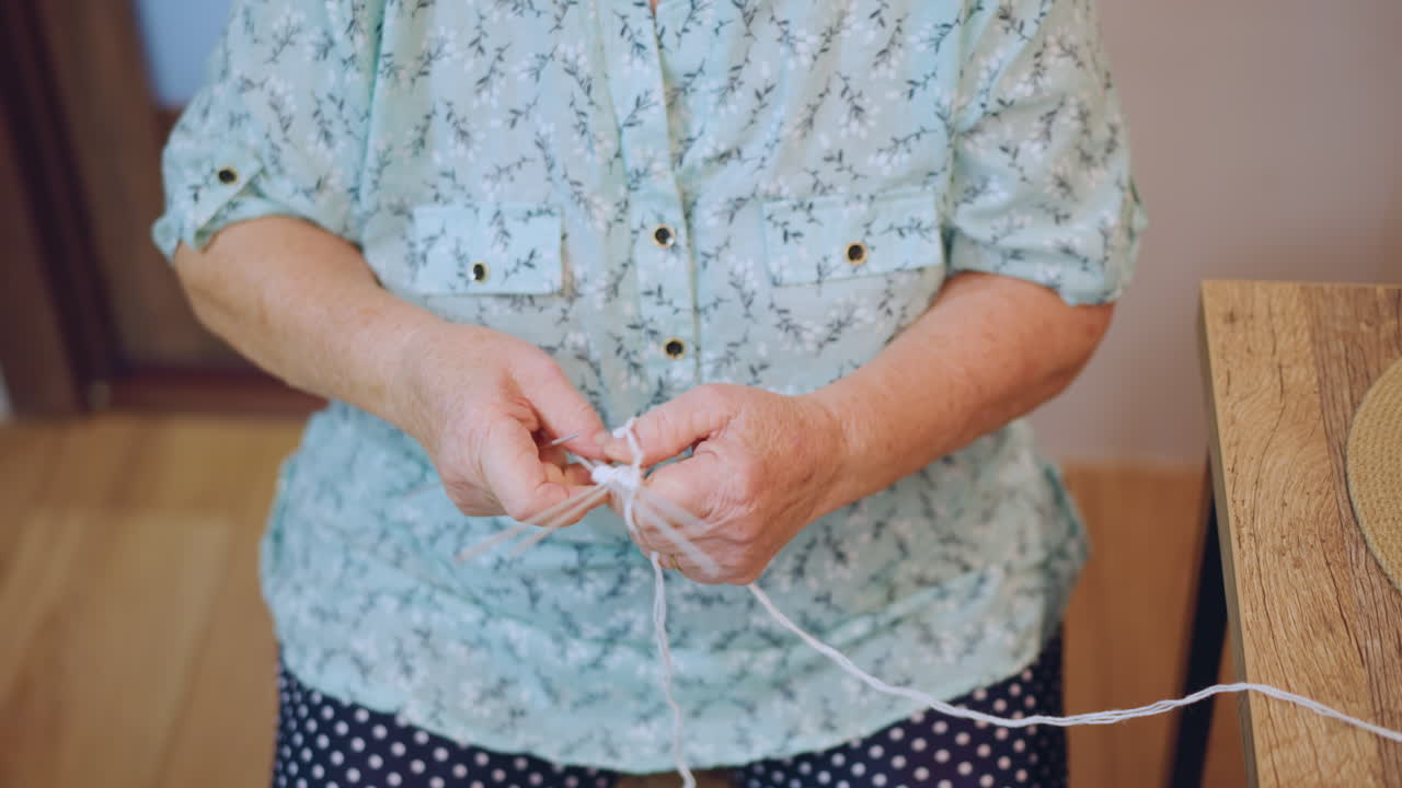Senior lady untangling colorful thread carefully. Old woman sitting at table with yarn and flowers. Elderly lady dressed in floral attire engaging in domestic craft activity indoors