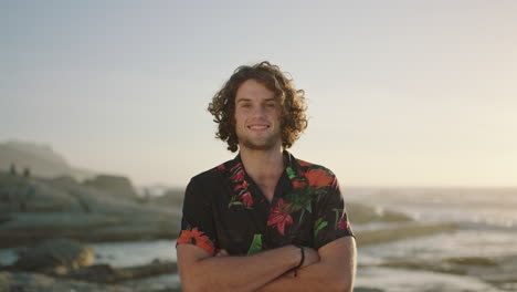 portrait of relaxed young man with arms crossed smiling at beach wearing hawaiian shirt