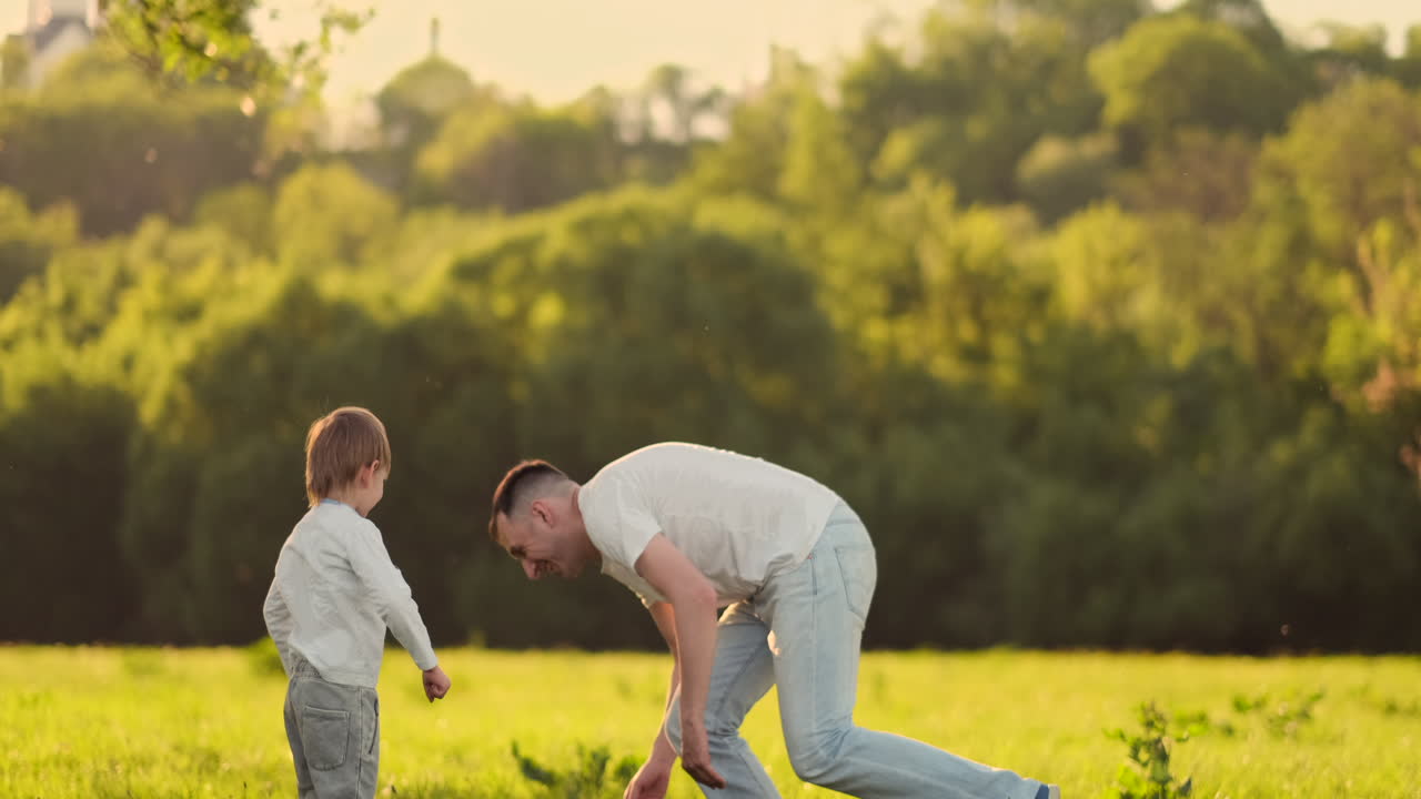 un joven padre en una camiseta blanca con dos hijos jugando al fútbol en el césped al atardecer en el sol en cámara lenta