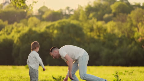 Un-Joven-Padre-Con-Una-Camiseta-Blanca-Con-Dos-Hijos-Jugando-Al-Fútbol-En-El-Césped-Al-Atardecer-Bajo-El-Sol-En-Cámara-Lenta