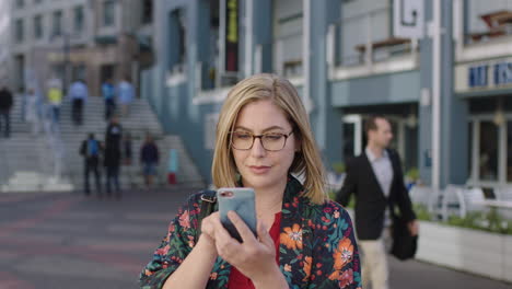 portrait-of-happy-young-blonde-woman-texting-browsing-using-smartphone-social-media-app-wearing-glasses-in-urban-background