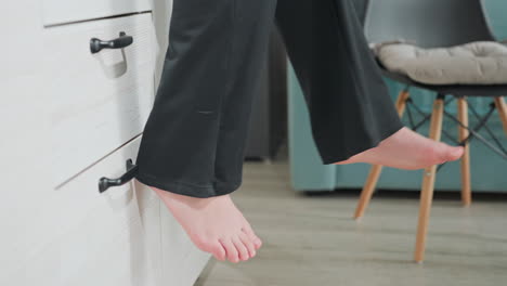 side view of adult legs swinging while seated on white wood cabinet edge in modern cozy kitchen interior with soft natural light illuminating motion and texture of black pants