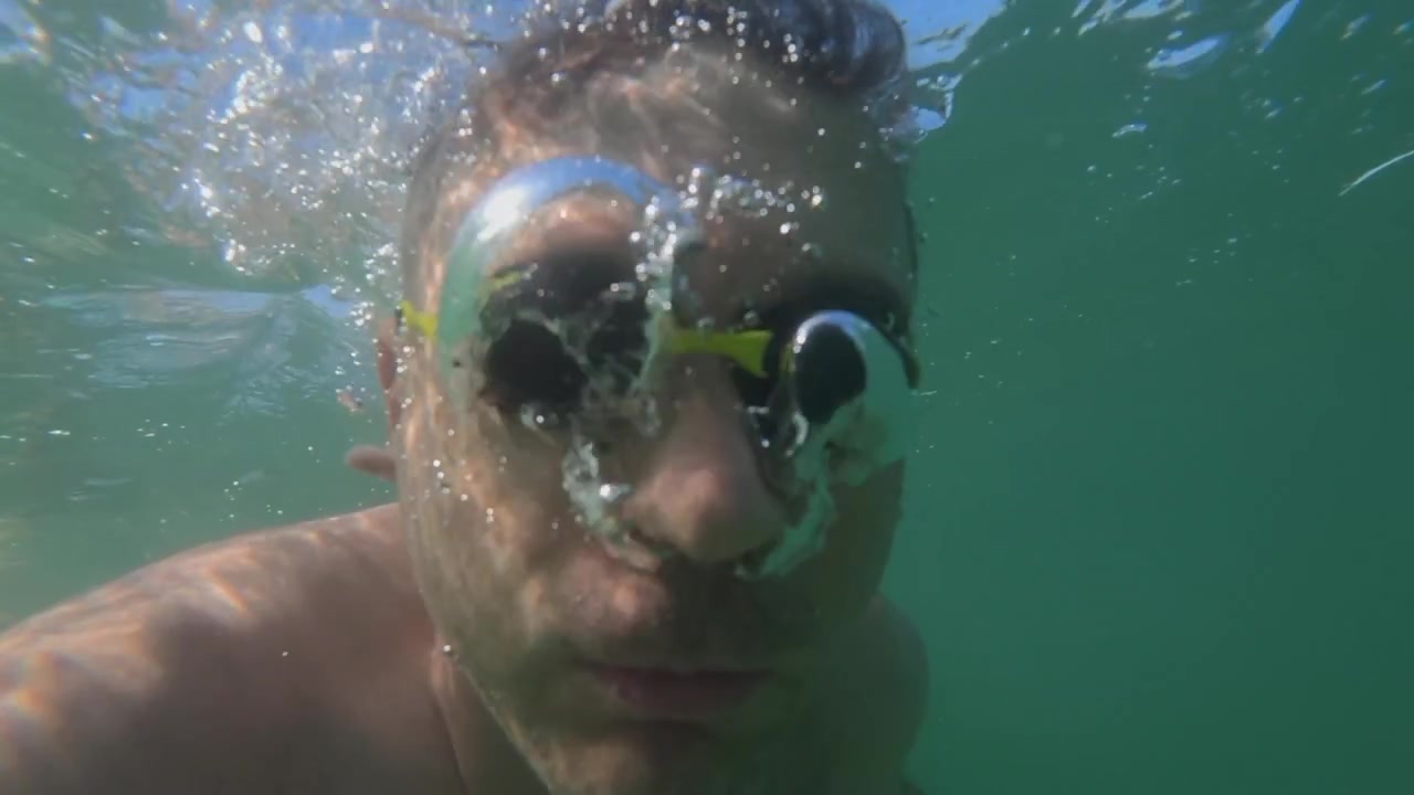 Persona che indossa occhialini da nuoto con il viso parzialmente sommerso, che guarda in su da sotto la superficie con bolle d'acqua.