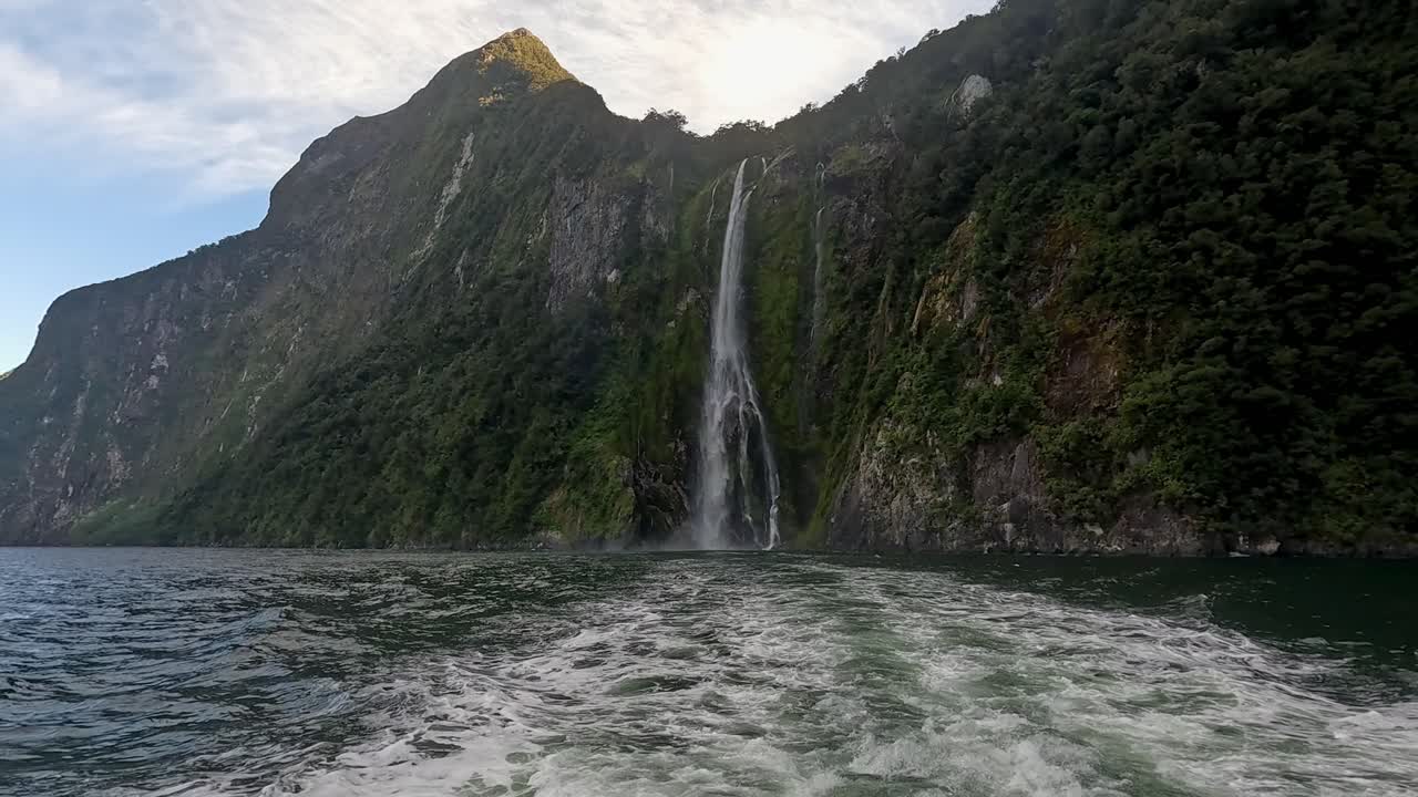 View from the back of a boat sailing away from a towering waterfall in Milford Sound, New Zealand. This is in the Fiordland National Park in New Zealand's South Island
