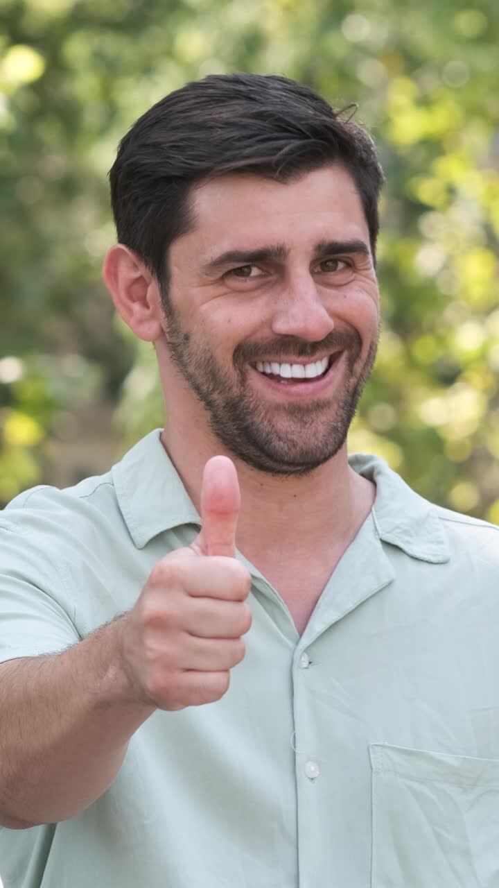 Uomo felice in camicia casual che mostra il pollice in su in un parco cittadino in una giornata di sole.