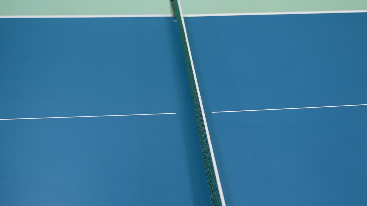 Aerial view of tennis ball flying above blue table court during match showing net centerline focus, precision play, competitive atmosphere, movement, and dynamic sporting action