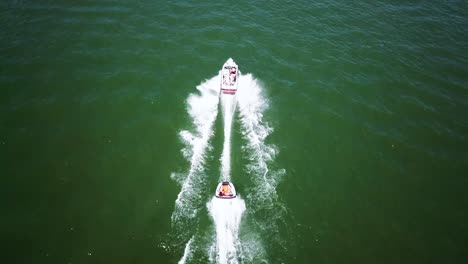 Drone-Shot-of-a-boat-on-a-green-lake-pulling-behind-it-a-tube-with-people-on-it,-creating-a-large-wake-area