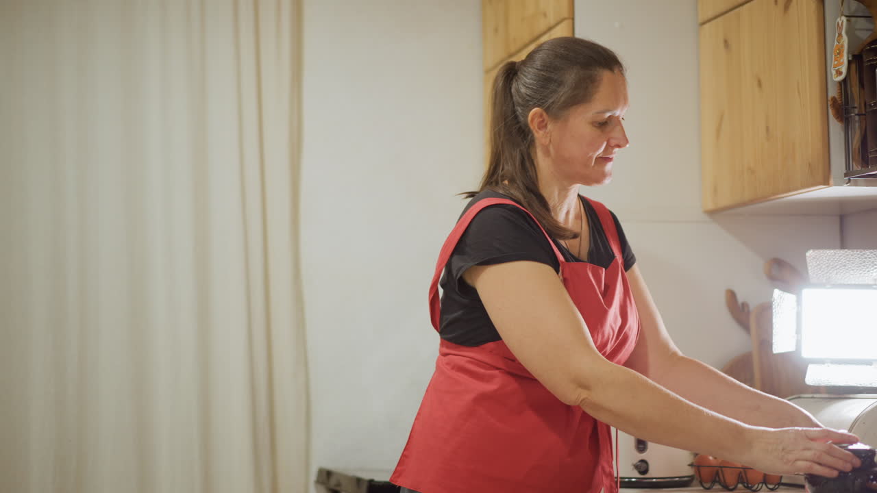 Femme blanche préchauffant le four dans sa cuisine. La cuisinière à domicile portant un tablier rouge vérifie le four, glisse un plateau de pâte dans le four, règle la minuterie, attrape un saladier et des œufs sur le comptoir, bouilloire et pot en céramique