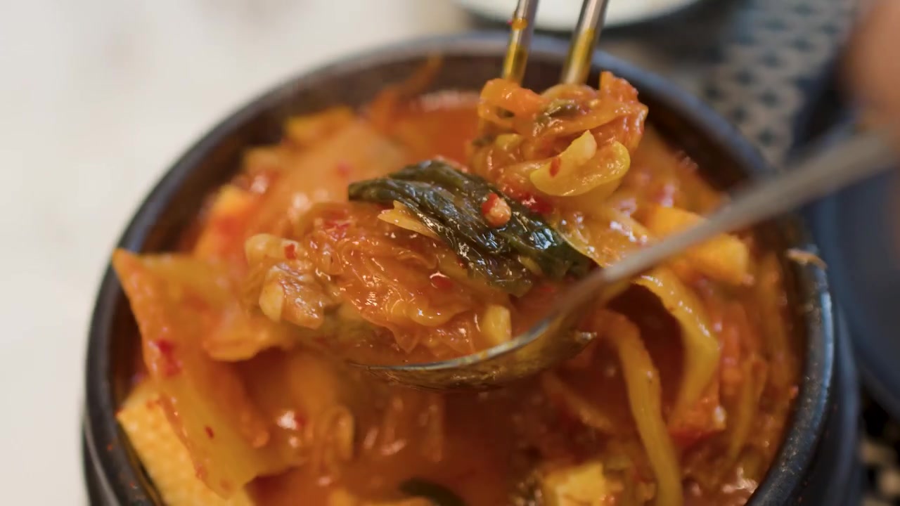 Tampilan close-up dari atas menunjukkan sendok logam mengaduk sup tahu kimchi pedas panas dalam dolsot.