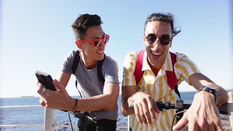 happy diverse gay male couple using smartphones at promenade by the sea, slow motion