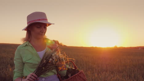 woman in a hat with a bouquet of wild flowers walking around the field at sunset 4k video