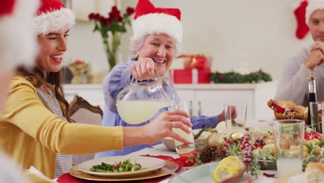 Kaukasische-ältere-Frau-Mit-Weihnachtsmütze-Gießt-Getränk-In-Ein-Glas-Einer-Frau,-Während-Sie-Am-Esstisch-Sitzt