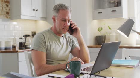 Happy-caucasian-man-sitting-at-table-in-kitchen,-using-laptop-and-talking-on-smartphone