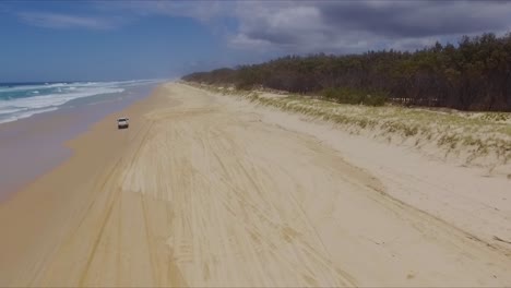 smooth, wide aerial drone shot following a white 4x4 four wheel drive pickup truck along north stradbroke island's main beach from a distance, as clouds gather over the island
