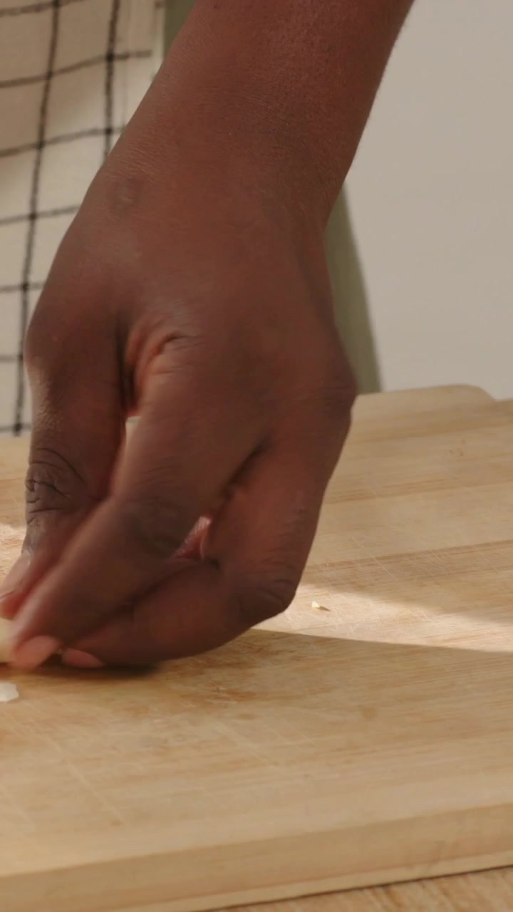 Video verticale: Mani di un cuoco che abbassano un coltello blu, tritando aglio su un tagliere in una ciotola di vetro.