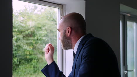 an anxious worried businessman is stressed with head in hands while he is looking out of a window at the rain