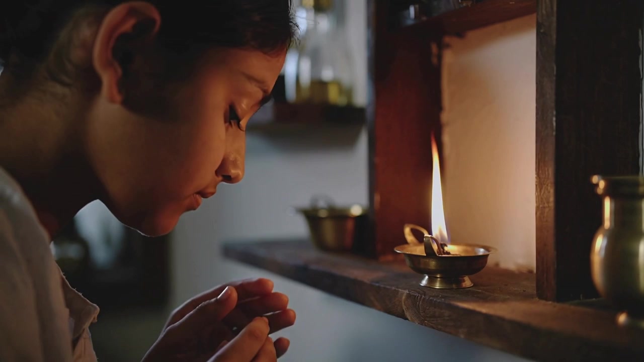 Bringing match, girl igniting diya in home shrine for prayer, wearing beaded bracelet, cupping hand
