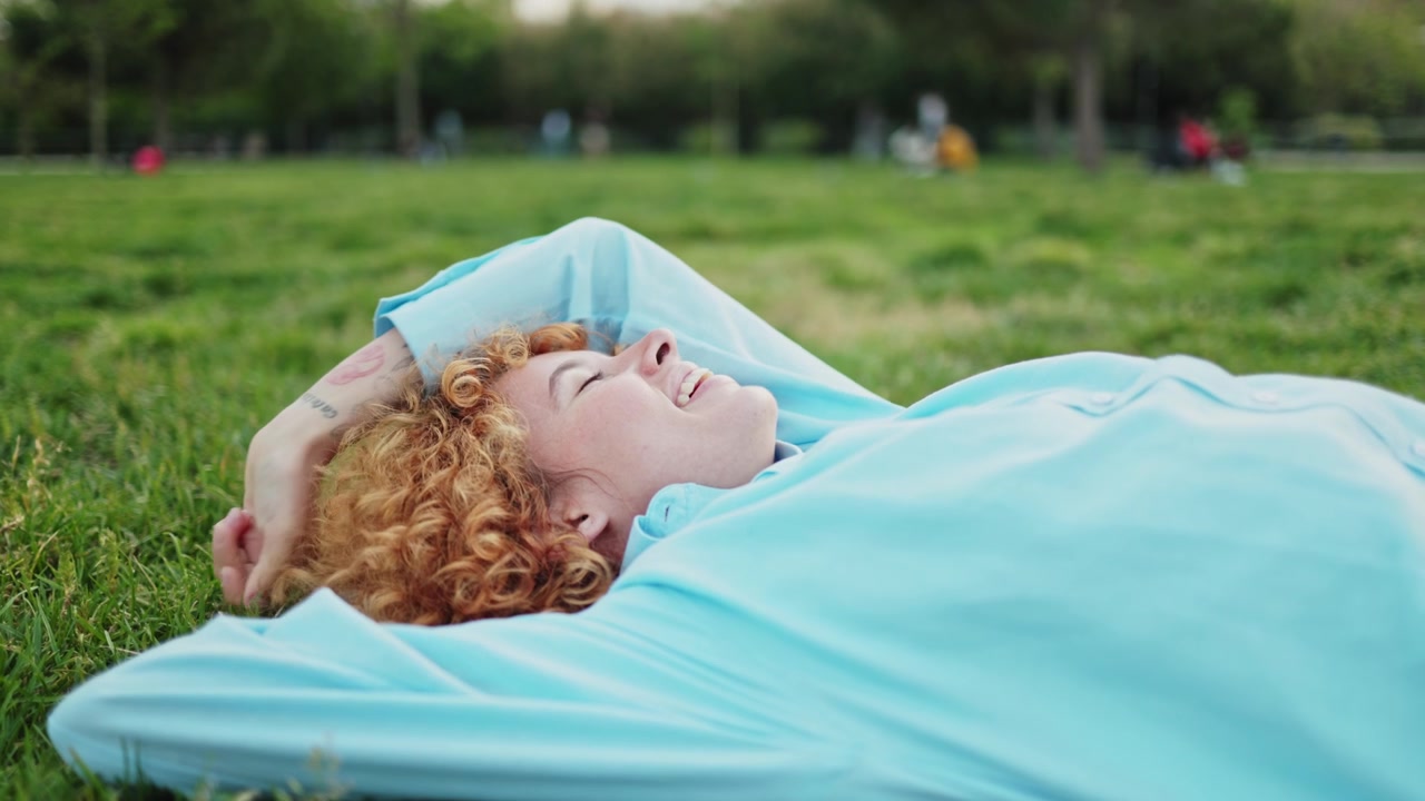 Peaceful Young Woman with Curly Red Hair Relaxing on Grass in Park