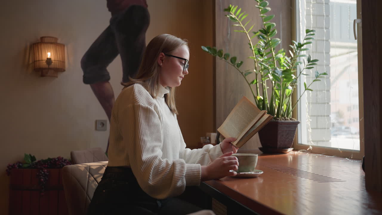 Side view of woman sitting by window reading book and holding coffee cup in warm cozy cafe with indoor plant, soft lighting, and calm ambiance during peaceful afternoon moment