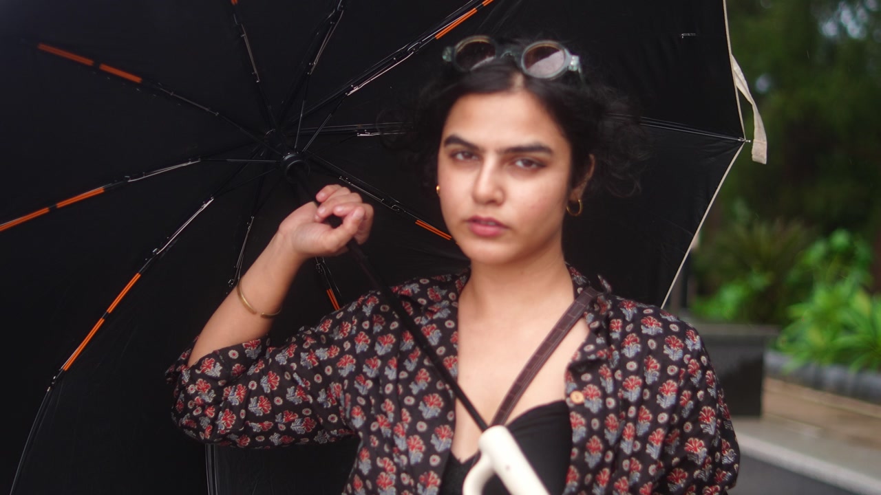 Stylish Indian woman sheltering under umbrella, wearing contemporary clothing amid verdant landscape during rainy season in Goa, western India