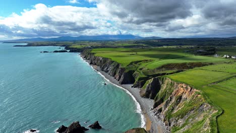 Drone-flying-along-dramatic-cliff-formations-emerald-green-seas-white-waves-mountains-in-background-and-puffy-clouds-Waterford-coast-Ireland-on-a-spring-morning