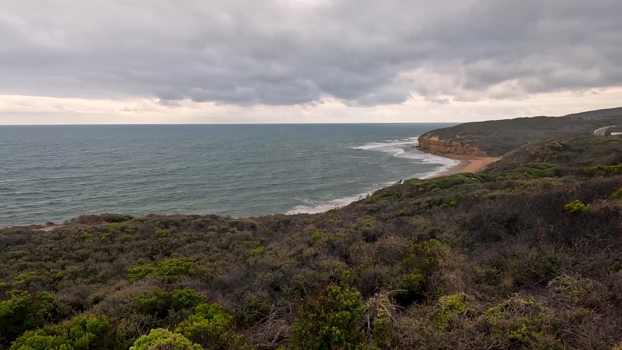 Aerial camera slowly pans across rugged coastal cliffs, dense green shrubland, and choppy ocean under overcast skies, capturing dramatic Great Ocean Road scenery
