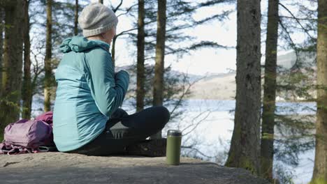 A-woman-sits-cross-legged-on-a-rock-eating-her-lunch-while-out-for-a-walk-in-the-forest,-looking-out-across-a-Scottish-Loch-and-hillside-in-the-sun