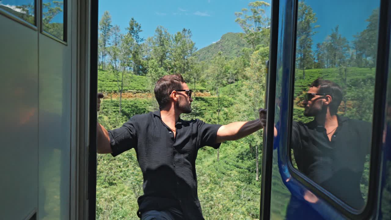 A man sits on the open door of a train traveling the scenic Ella-Kandy route in Sri Lanka. Behind him, expansive tea plantations spread across rolling hills, showcasing the region’s iconic landscape.