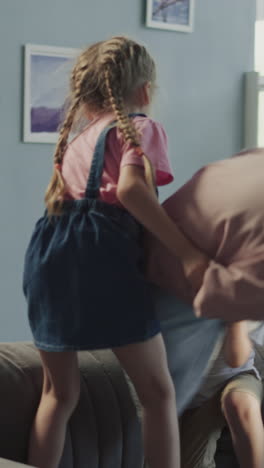 happy elder sister and brother have pillow fight standing on grey sofa at home. delighted little boy and girl play actively together in living room