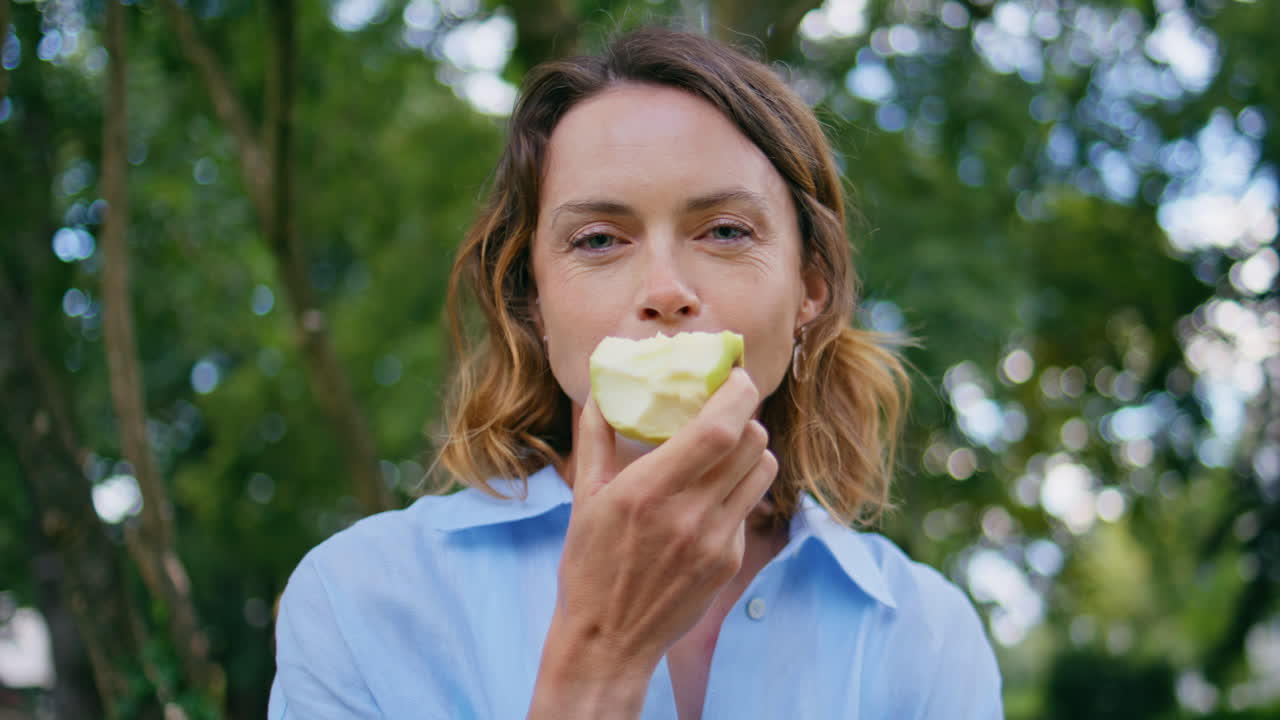 Happy woman eating fruit at park portrait. Lady enjoying apple looking camera