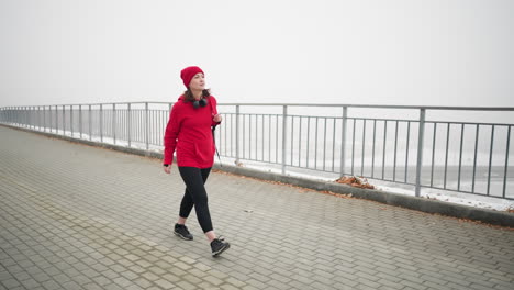 freelancer en chaqueta roja de invierno ajusta la bolsa sobre el hombro mientras camina a lo largo de un camino entrelazado cerca de la barandilla de hierro con follaje seco esparcido, atmósfera brumosa y coche lejano visible debajo del puente