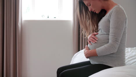 depressed pregnant woman sitting on bed holding belly