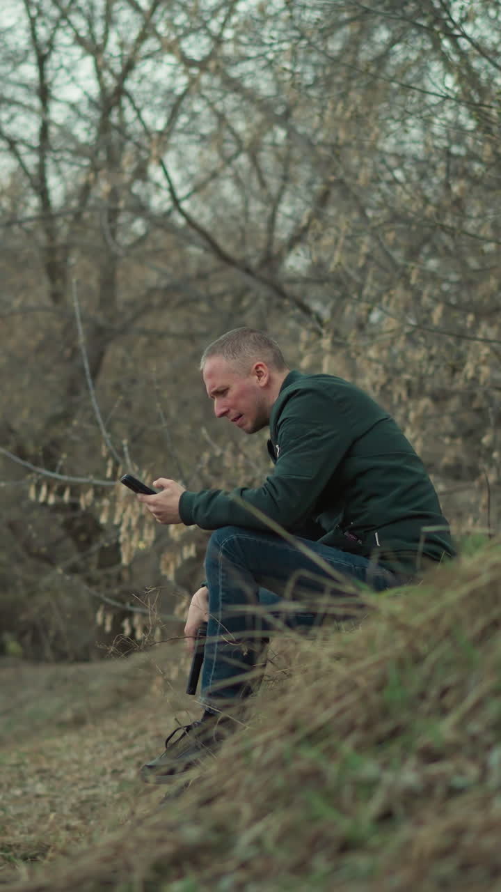 A close view of a man sitting on a grassy hillside with bare trees in the background, holding a gun and looking down pensively as he operate his phone