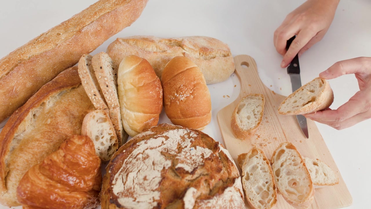 Overhead shot of hands slicing a baguette on a board surrounded by artisan breads.